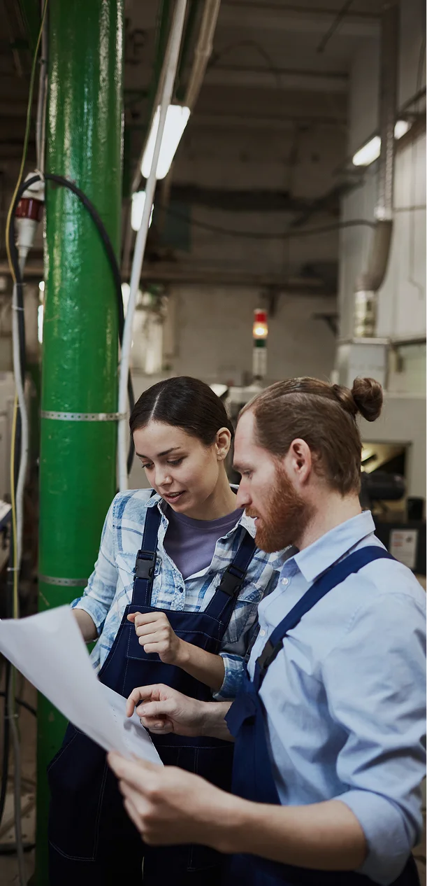Two colleagues in blue overalls review a document in an industrial plant by large green machinery.