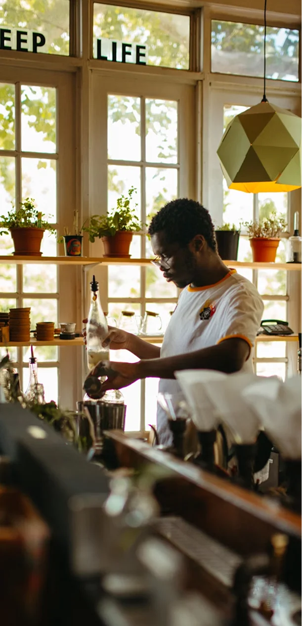 A man behind a bar bottle-service, pouring from a bottle into a shaker in a sunlit cafe with plants on the shelves.