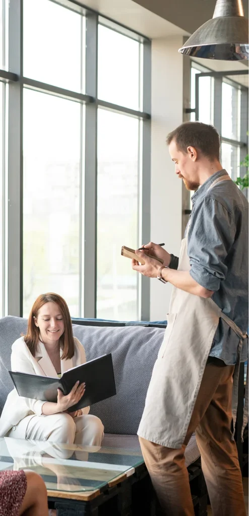Waiter in an apron stands by a couch, showing a notebook to a seated woman who is reading a menu in a bright cafe.